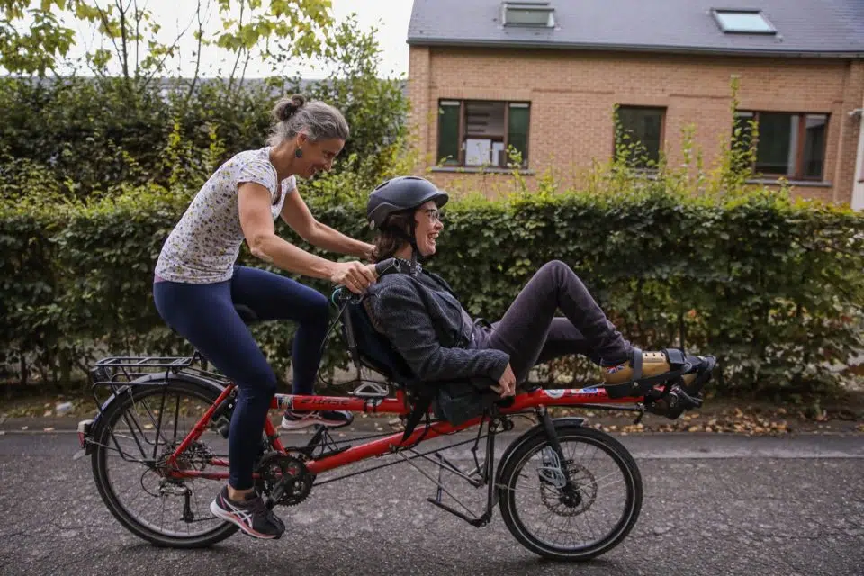 Deux membres de l'Asbl Escalpade : une personne handicapée et son accompagnatrice pédalant sur un tandem