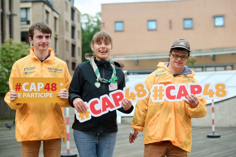 Trois bénévoles CAP48 montrent en souriant des panneaux #CAP48 à la caméra.